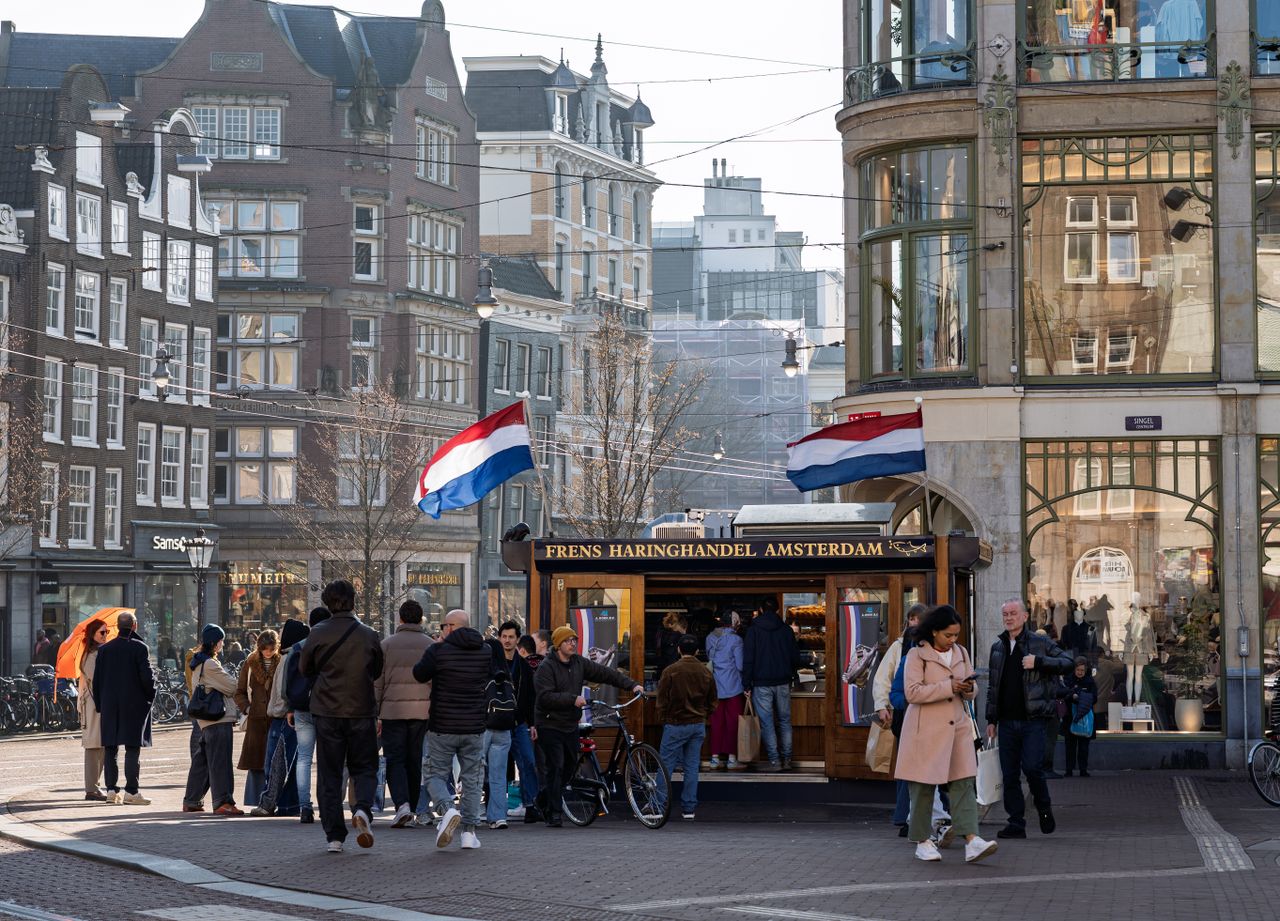 A lively Amsterdam street with people near a traditional Dutch herring stall, flags waving.